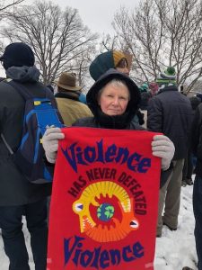 A woman in a heavy black jacket and wool gloves stands in a crowd with snow on the ground holding a red banner that says "violence has never defeated violence"