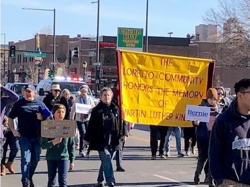 The street view of a march for justice with numerous people. The large yellow banner focused on in the picture states: The Loretto Community honors the memory of Martin Luther King Jr.