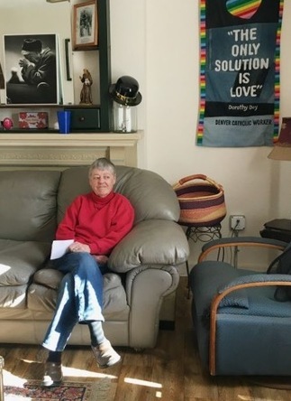A woman with short grey hair, wearing a red sweater and blue jeans sitting on a couch in a living room looking out the window candidly.