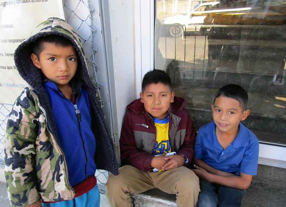 Three young boys with short dark hair, two sitting and one standing together looking at the camera for a picture. They are outdoors next to a wire fence in front of a glass window.