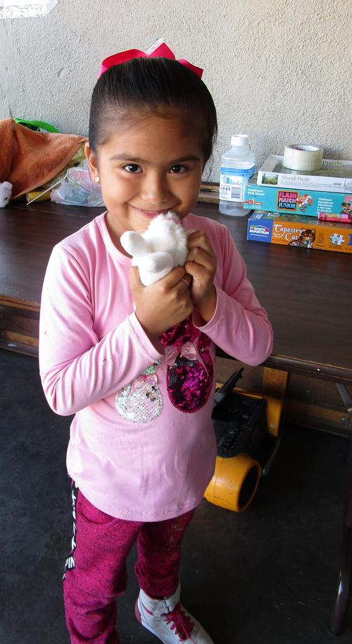 A young girl wearing a light pink shirt, a dark pink bow, and dark pink pants smiling brightly while holding a small white stuffed animal in her hands.