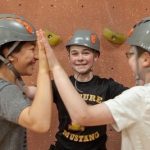 Three friends high-fiving in a circle while indoor rock climbing wearing grey helmets.