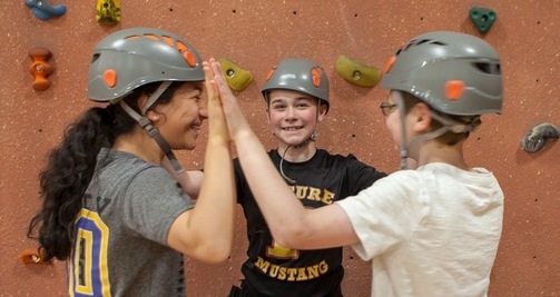 Three friends high-fiving in a circle while indoor rock climbing wearing grey helmets.