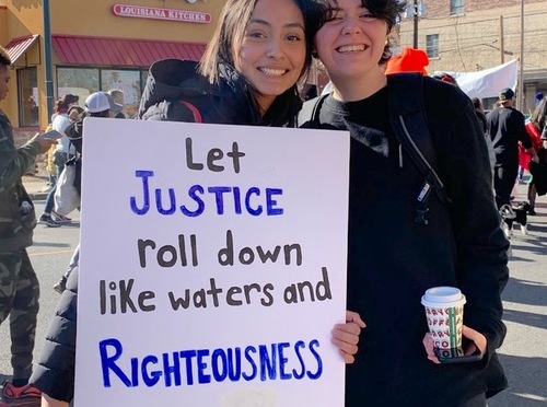Two young women smile together while one holds a sign stating: Let justice roll down like waters and righteousness. They are attending an outdoor march with numerous people behind them.