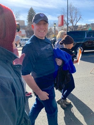 A man wearing a baseball cap holding a jacket in his arm smiles brightly for a picture while walking with others outdoors on a sunny day.