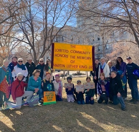 A large group picture outdoors on a sunny day. in the back center of a group, they are holding a large yellow sign that states: The Loretto Community honors the memory of Martin Luther King Jr.