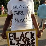 A black woman wearing a covid mask and a white shirt that says: Black girl magic holding a protesting sign at an outdoor protesting event. Her sight says: Black Lives Matter