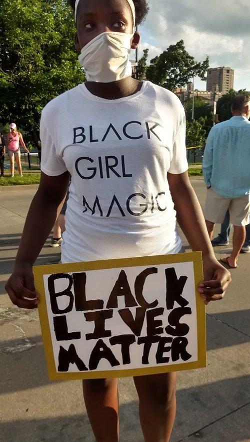 A black woman wearing a covid mask and a white shirt that says: Black girl magic holding a protesting sign at an outdoor protesting event. Her sight says: Black Lives Matter