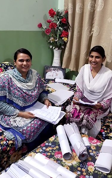 Two women, wearing long-sleeve patterned dresses smiling together while sitting in a living room sorting papers together.