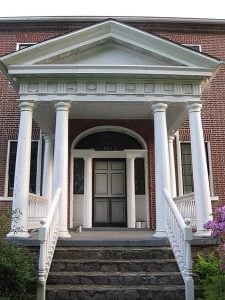 The front entrance of a red brick building with a large white awning with 4 columns creates a grand entrance up the stairs for the front door.
