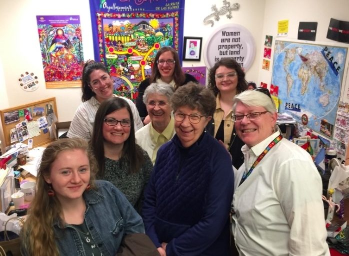 Eight women smiling together for a group picture in a small room with maps, art, and posters decorating the walls.
