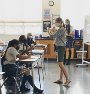 A teacher standing at the front of a classroom talking to students sitting at their desks. They are all wearing COVID masks.