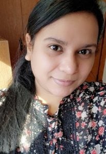A woman taking a selfie, she has dark brown long hair pulled back in a ponytail and she is wearing a black and floral patterned blouse.