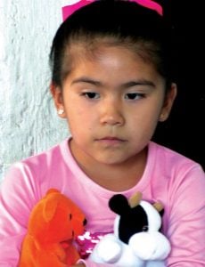 A child with black hair tied in a pink bow wears a pink shirt and is holding two stuffed animals looking past the camera.
