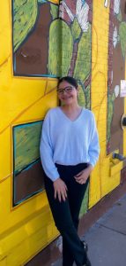 A woman with dark brown hair and dark-framed glasses smiling brightly wearing black pants, black boots, and a white sweater while posing standing next to a building mural of big yellow letters with cactuses in the background.