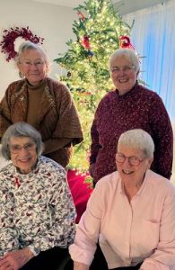 Four women, two sitting and two standing smiling together for a group picture indoors in front of a lit Christmas tree.