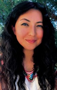 A headshot of a woman with long wavy black hair smiles softly. She is wearing a white shirt with a detailed colorful embroidered pattern at the top.