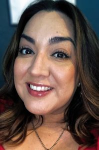 Close up headshot of a woman with dark brown hair with highlights smiling. She is wearing black eyeliner, black mascara, a red lip gloss and a silver chain necklace.