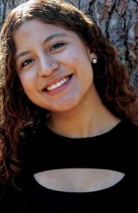 A woman with wavy brown hair, chunky stud earrings and a black shirt smiling.