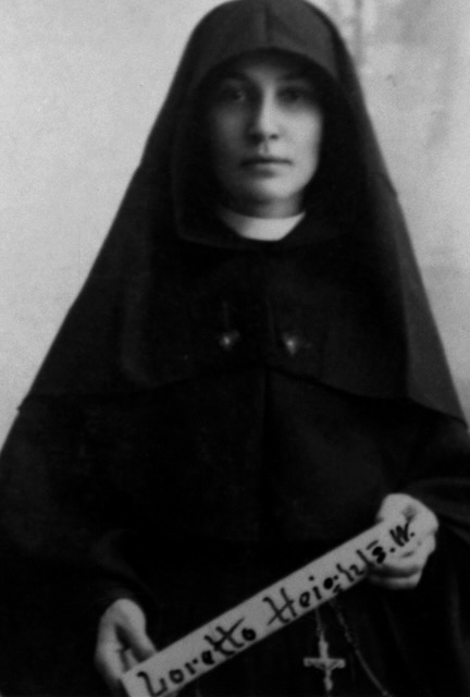 A blurry black and white photo of a religious sister in a habit holding a skinny sign that says "Loretto Heights."