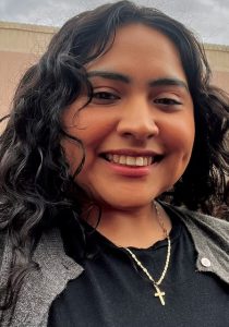 A woman with curly black hair smiles for a selfie. She is wearing a black shirt, grey cardigan and a gold cross necklace.