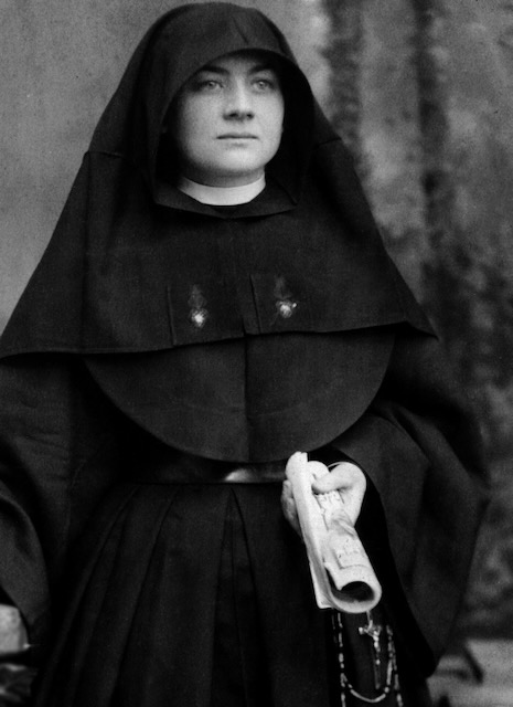 A black and white photo of a nun in habit looks off to the side and is holding a rolled up book.