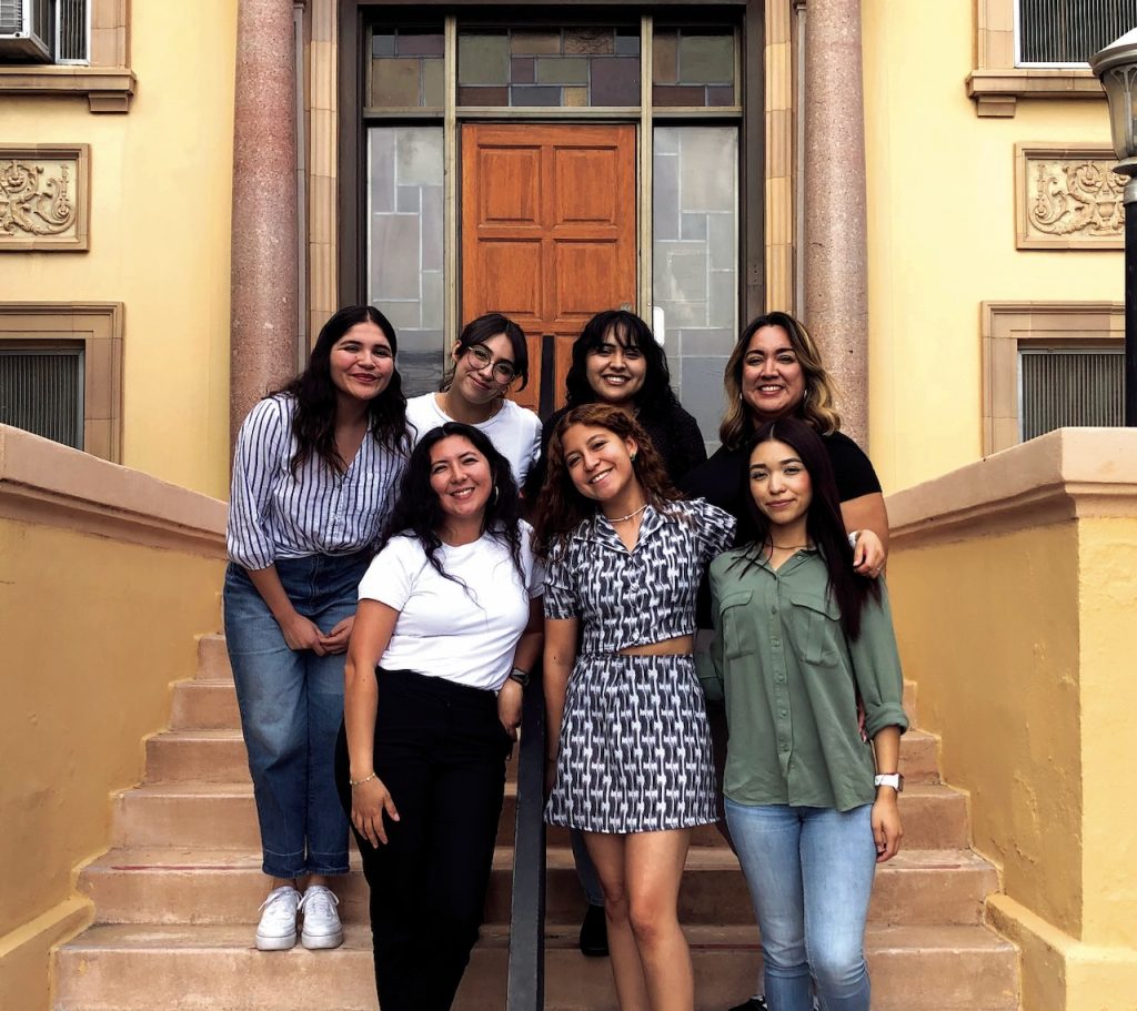 Seven woman stand for a group photo staggered on clay steps in front of a building with four in the back and three in the front.