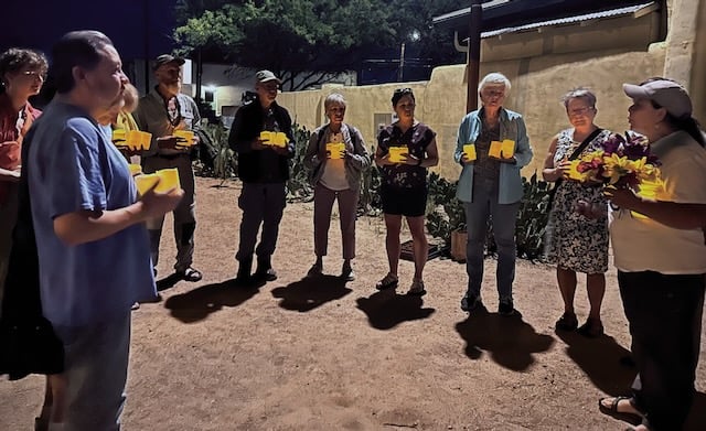 Eleven people stand in a semi-circle in a sandy area holding candles for a vigil.