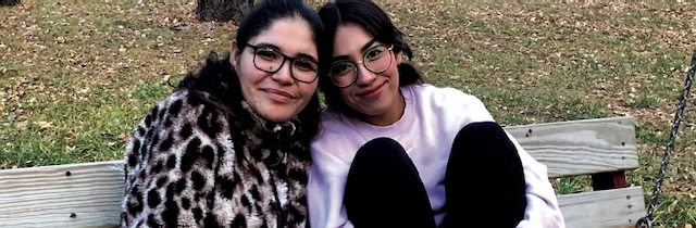 Two women with glasses and dark brown hair sit close together on a wooden bench as they smile softly for the camera.