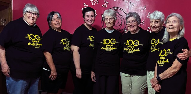 A group of seven women standing in a row smiling in front of a red wall wearing matching black t-shirts with yellow print that says"100 years of Sisterhood, Loretto Academy."