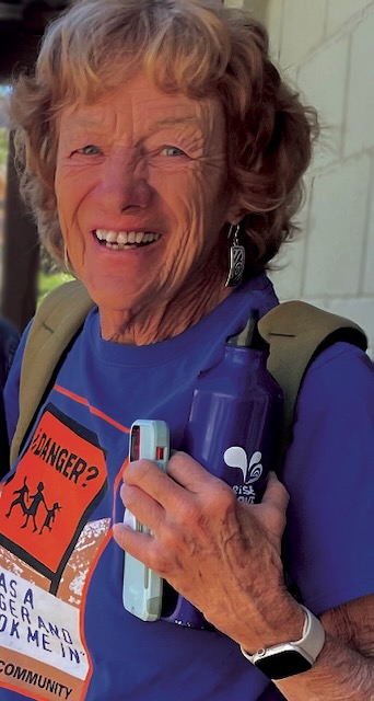 Photo of an older woman smiling. She has dark blonde short curly hair, dangly silver earrings, is wearing a purple shirt, green backpack and holding a purple water bottle and light blue iPhone.
