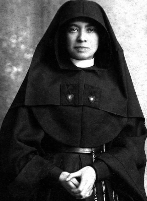 A religious sister standing wearing a habit and looking directly at the camera with her hands held together.