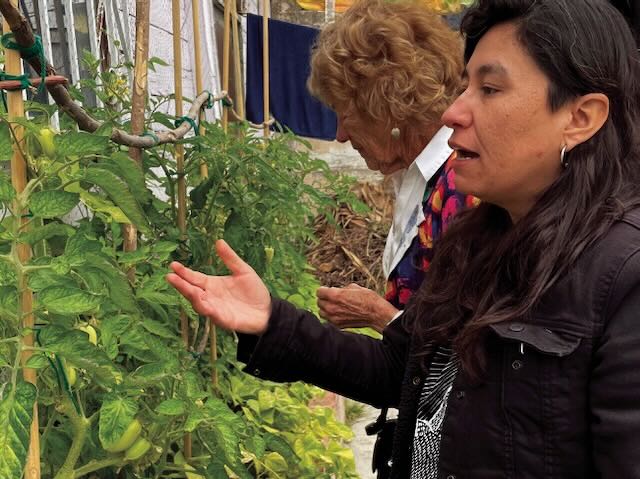 A woman in a black jacket gestures towards plants in a garden as she gives a tour. A woman behind her is seen looking at the plants.