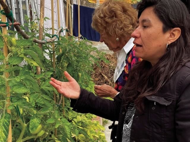 A woman in a black jacket gestures towards plants in a garden as she gives a tour. A woman behind her is seen looking at the plants.