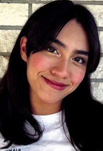 A woman with black hair and a white shirt smiles softly. She is wearing gold eyeliner, black mascara and reddish pink lipstick.