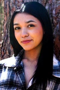 A woman with long black hair and a plaid button up shirt smiles softly and looks directly at the camera.