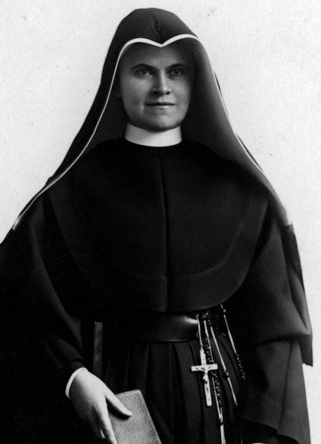 A religious sister in a habit and robes is standing looking past the camera holding a small book.