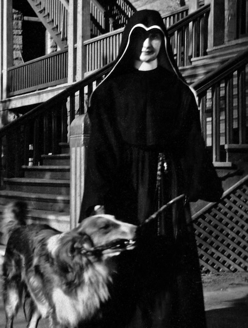 A religious sister in a habit stands in front of a large staircase with a dog near her side.