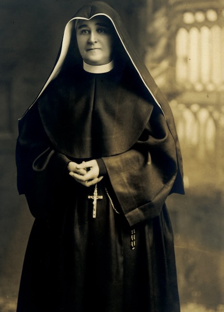 A sepia toned photo of a religious sister in a black habit and robes standing and holding a rosary.