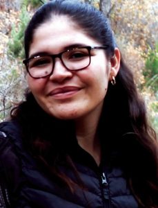 A woman with long brown wavy hair smiles without teeth for a headshot. She is wearing square brown glasses, small gold hoop earrings and a blue puffy jacket.