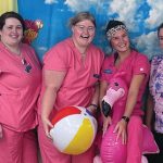Eight smiling women of a variety of racial backgrounds wearing pink and colorful nursing scrubs pose in front of a beach background with a beachball and inflatable flamingo as props.