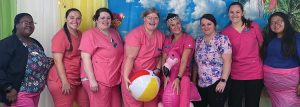 Eight smiling women of a variety of racial backgrounds wearing pink and colorful nursing scrubs pose in front of a beach background with a beachball and inflatable flamingo as props.