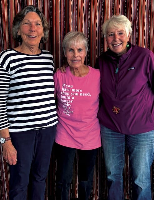 Three women stand side by side smiling in front of a striped vertical patterned backdrop.