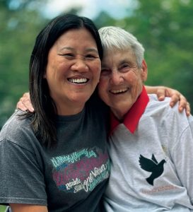 Two woman hold each other close side by side and smile in a photo. The woman on the left has longer black hair and is wearing a graphic t-shirt. The woman on the right is older and has short grey hair, wearing a grey sweatshirt over a red collard shirt.  