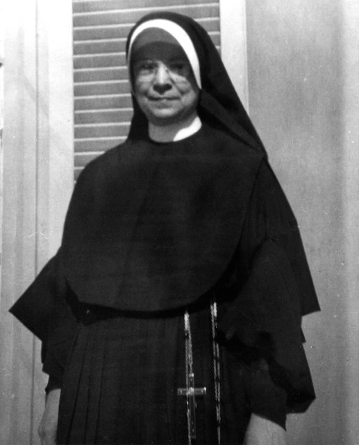A black and white photo of a nun in habit standing looking at the camera. She is wearing clear framed rectangular glasses and a rosary hangs from her waist.