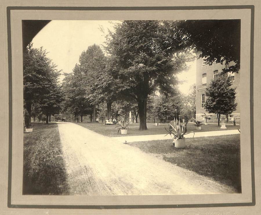 Archival photo in sepia tones of a tree-lined road in front of a three-story building.
