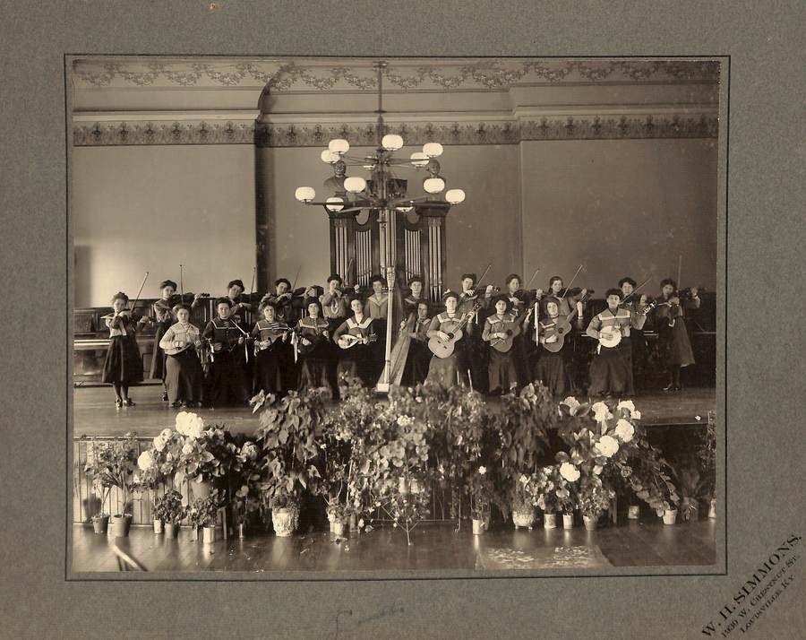 Archival photo in black and white of student musicians on a stage with a great number of potted plants of all shapes and sizes lining the floor in front of the stage.