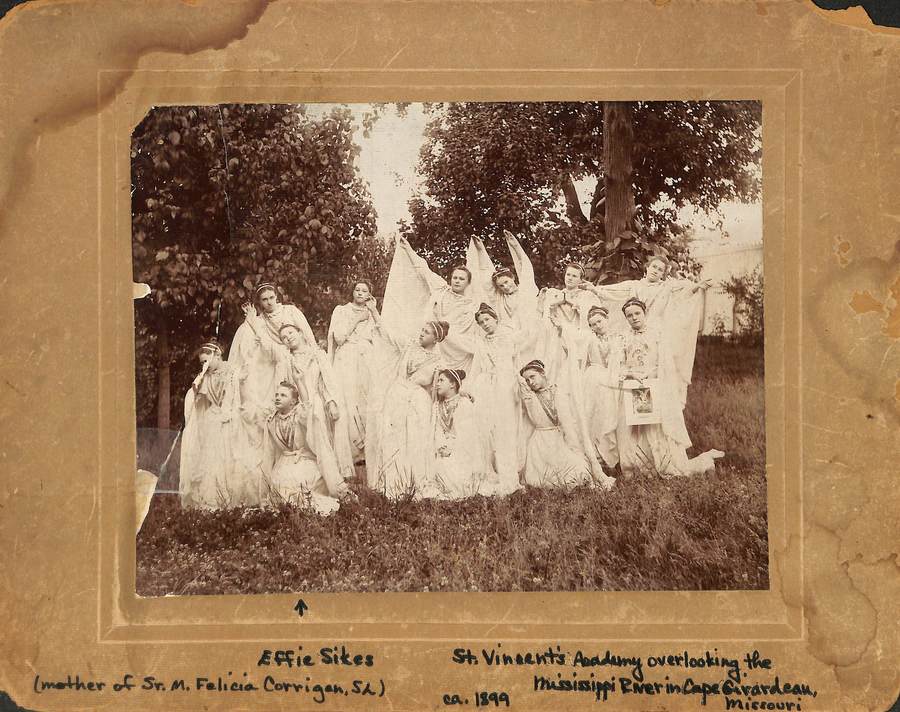 Archival photo in sepia tones of a group of women in white dresses posing dramatically for the photo. Handwritten text on the mat of the photo reads "St. Vincent's Acadamy overlooking the Mississippi River in Cape Girardeau, Missouri, ca. 1899" An arrow points up to one of the women with the text "Effie Sikes "mother of Sr. M. Felicia Corrigan, SL)"
