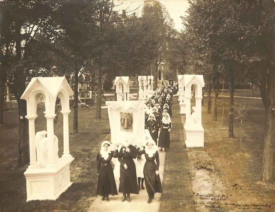 Archival photo in sepia tones of a procession of sister in habit carrying a banner down a path from the Loretto Motherhouse church. Handwritten text on the photo reads "'Foundation Day' April 25, 1912"
