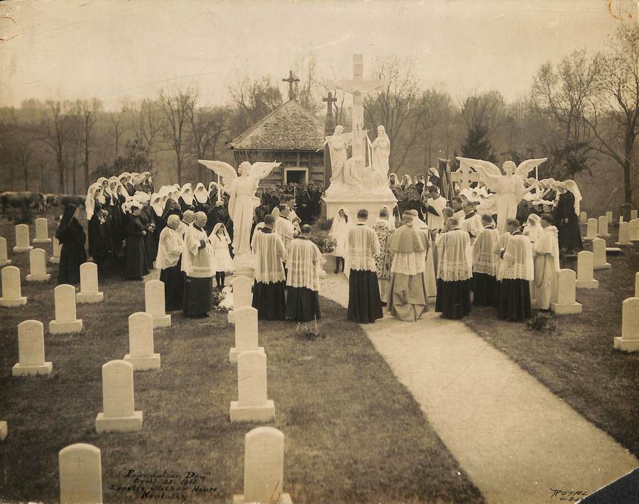 Archival photo in sepia tones of a photo of religious gathered in a circle for an event in the Loretto Motherhouse cemetery. Multiple rows of sisters in habit stand at the back, with clergy in full cassocks and accoutrements in the foreground. All are facing the large statue of the crucifixion in th center.
